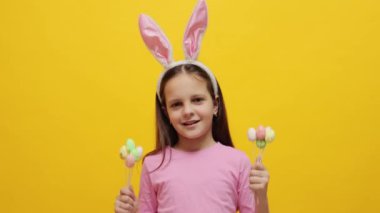 Gülümseyen küçük kız pembe tavşan kulaklığı takıyor elinde pop kekler sarı arka planda izole edilmiş lezzetli şekerlerle mutlu tatiller kutluyor.
