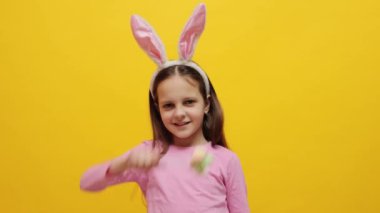 Son derece mutlu küçük bir kız pembe tavşan kulaklığı takıyor elinde pop kekleri var sarı arka planda izole bir şekilde duruyor bahar tatilini kutlayan şekerlerle dans ediyor.