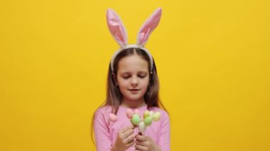 Pembe tavşan kulaklığı takan, sarı arka planda izole bir şekilde duran ve şekerlerini yiyen parti için hazırlayan küçük bir kız.