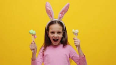 Çocukların neşeli Paskalya kutlaması. Aşırı neşeli iyimser küçük kız pembe tavşan kulaklığı takıyor elinde pop kekler sarı arka planda izole edilmiş çığlıklar atıyor heyecanla haykırıyor