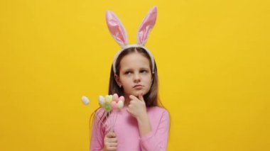 Paskalya kutlaması. Düşünceli küçük bir kız, tavşan kulaklığı takıyor, elinde pop kekler, sarı arka planda izole bir şekilde duruyor, çenesini kapatmış, düşünceli bir yüzle Paskalya kutlamasını düşünüyor.