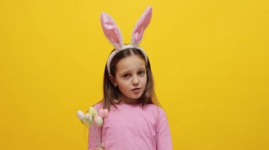 Küçük bir kızın Paskalya kutlaması. Pembe tavşan kulaklığı takan kendine güvenen küçük kız çocuğu sarı arka planda izole bir şekilde duran pop kekleri parmağıyla inkar hareketini gösteriyor.