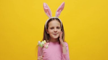 Heyecanlı küçük kız pembe tavşan kulaklığı takıyor elinde pop kekleri var sarı arka planda izole bir şekilde duruyor çığlık çığlığa güve sesleri çıkarıyor.