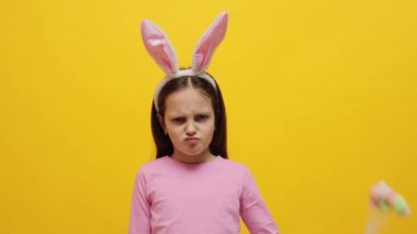Kızgın küçük kız pembe tavşan kulaklığı takıyor elinde pop kekleri var sarı arka planda izole edilmiş poz veriyor ve hayır diyor parmak hareketleriyle nefret ve anlaşmazlıkla