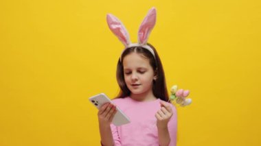 Bahar kutlamaları için tavşan temalı dekorasyonlar. Küçük kız pembe tavşan kulaklığı takıyor elinde pop kekler var sarı arka planda izole bir şekilde duruyor cep telefonuyla internette geziniyor.