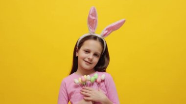 Paskalyayı çocuk partisiyle kutluyoruz. Pembe tavşan kulaklığı takan sevimli küçük kız sarı arka planda izole edilmiş pop kekleri tutuyor tatlı tebessümleriyle sarılıyor
