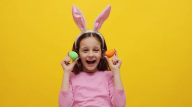 Çocuklar için Paskalya kutlaması. Sarı arka planda izole edilmiş renkli paskalya yumurtalarını tutan pembe tavşan kulaklığı takan şenlikli küçük kız heyecandan bağırıyor.