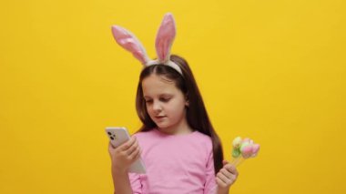 Pembe tavşan kulaklığı takan sevimli küçük kız elinde pop kekleri tutarak sarı arka planda izole bir şekilde poz veriyor elinde akıllı telefon internet sayfaları tutuyor.