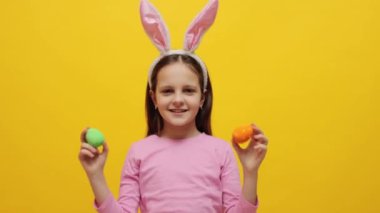 Çocuklar için Paskalya yumurtası avı heyecanı. Pembe tavşan kulaklı mutlu küçük kız elinde renkli Paskalya yumurtaları, sarı arka planda, bahar partisini kutluyor.