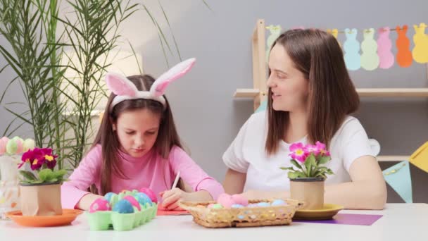 イースターのお祝いのためのグリーティングカードと絵画の卵を描く親切な家族の雰囲気でテーブルに座っている母親を持つ小さな少女 — ストック動画