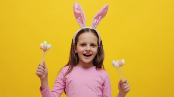 ピンクのウサギの耳を身に着けている小さな女の子を踊る 黄色の背景に隔離されたポップケーキを抱く 美味しいデザートでイースターパーティーを楽しんでいます — ストック動画
