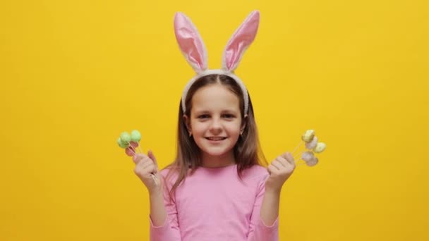 子供のためのウサギテーマのイースターパーティー デザートと笑顔で目を覆う黄色の背景の上にポップケーキを握っているピンクのウサギの耳を身に着けている喜ばしい愛らしい少女 — ストック動画