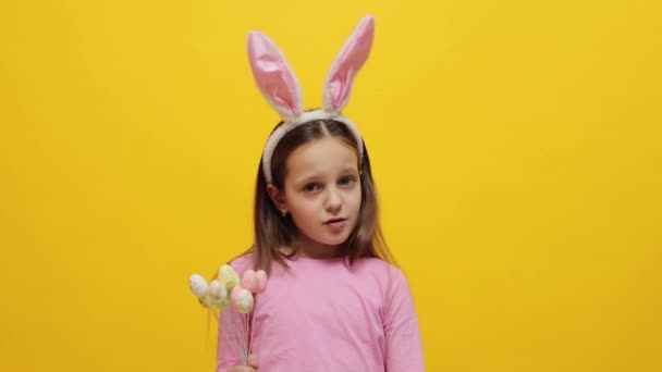 小さな女の子のイースターのお祝い 自信を持ってピンクのバニーの耳のヘッドバンドを身に着けている小さな女性の子供は 彼女の指で否定的なジェスチャーを示す黄色の背景の上に隔離されたポップケーキを保持しています — ストック動画
