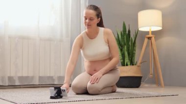 Çekici hamile kadın bej bluz giyiyor ve evde video eğitmeniyle egzersiz yapıyor telefon ve tripod kullanarak açık oturma odasının iç kısmında online egzersiz yapıyor.