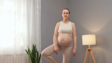Hamile kadın oturma odasında yoga yapıyor bej bluz giyiyor ve tayt giyiyor meditasyon yapıyor hamilelik sırasında zihin ve beden sağlığıyla ilgileniyor.