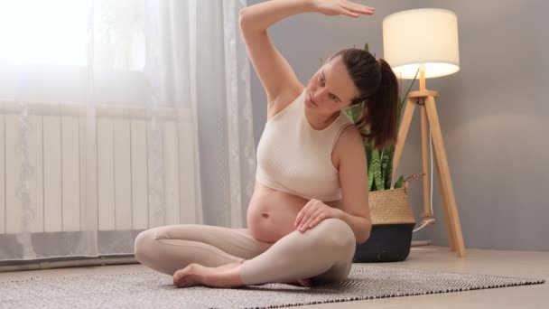 怀孕期间的瑜伽促进产妇健康和福祉 穿着米黄色上衣和腿的胖胖的孕妇在轻便的客厅里锻炼 — 图库视频影像