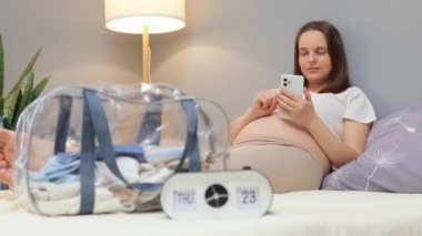 Elinde akıllı telefonuyla kahverengi saçlı hamile bir kadın bebek elbiseleriyle yatakta yatıyor. Arka planda doğum hakkında bilgi topluyor.