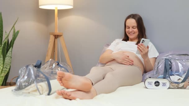 満足したかわいい魅力的な妊婦は 妊娠の最後の日を楽しんでいる幸せな表情で彼女の裸の腹をストロークで携帯電話でベッドで休む — ストック動画