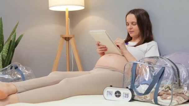 出産準備について 穏やかな茶色の毛髪の白人妊婦 ベッドに横たわる マタニティバッグのライティングチェックリスト 必要なものと項目をチェック — ストック動画