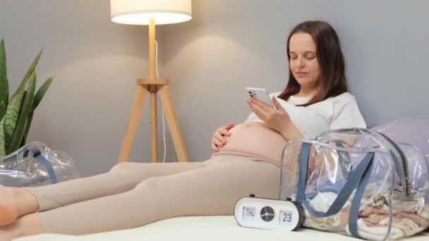 モバイルアプリケーションを使用して収縮間隔をチェックする産科病院に残る待っている携帯電話のブラウジングインターネットウェブページを使用して裸の腹を持つ好ましい妊婦 — ストック動画
