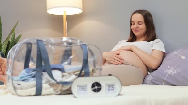 Nöjd Brunhårig Ung Gravid Kvinna Som Ligger Sängen Med Babykläder — Stockvideo