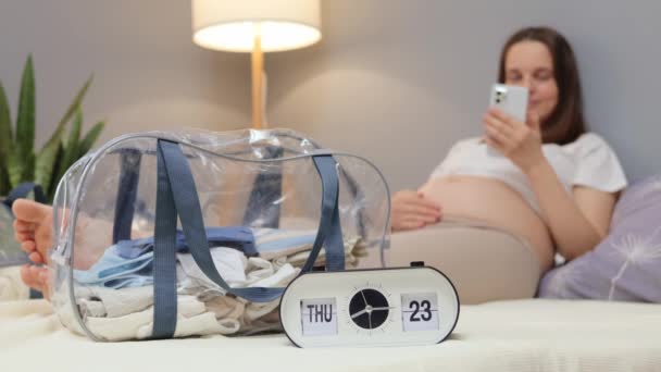 インターネットで出産に関する情報を読むバックグラウンドのバックグラウンドでベッドでベッドに横たわるスマートフォンを持つ若い妊婦との時計のクローズアップ — ストック動画