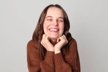 Kahverengi kazak giyen neşeli, kahverengi saçlı kadın açık gri arka planda tek başına durmuş mutluluğu ifade eden keyifli bir şey söylüyor.