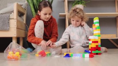 Küçük bebek kız ve ablası renkli yapı bloklarıyla oynuyorlar. Birlikte çalıştıktan sonra renkli detayları topluyorlar.