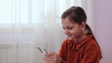 Mobil uygulama eğlencesi. İçeride ekran zamanı. Çocuk videoları. Mutlu küçük kız yatakta oturmuş yatak odasında akıllı telefonuyla arkadaşlarıyla sohbet ediyor.
