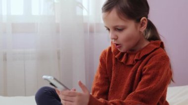 Çocukların dijital dünyası. Akıllı telefon keyfi için. Şaşırmış küçük kız yatak odasında cep telefonu kullanarak yatakta oturuyor ve internetteki şok olmuş çocukları buluyor.