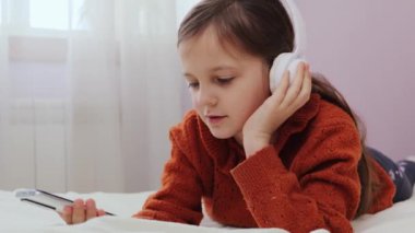 Yatak odasında kulaklıkla müzik dinleyen sevimli küçük kız elinde cep telefonu internet ortamında şarkı seçen favori parçanın tadını çıkarıyor.