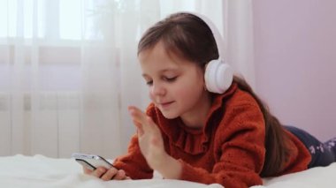 Kulaklıklı sevimli küçük kız yatak odasında uzanmış elinde cep telefonuyla video görüşmesi yapıyor ve arkadaşıyla el sallayarak selamlama hareketi yapıyor.