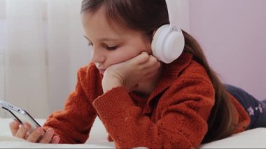 Sakin, kahverengi saçlı, kulaklıklı, akıllı telefon kullanarak yatakta uzanan sosyal ağlardaki web sayfalarını okuyan, çevrimiçi kaydırma yapmaktan hoşlanan küçük kız.