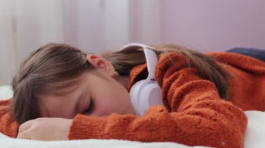 Kahverengi saçlı küçük bir kız kablosuz kulaklıklı bir çocukla yatağa uzanmış teknolojiyi kullanıyor online eğitim sırasında uykuya dalıyor veya müzik dinliyor.