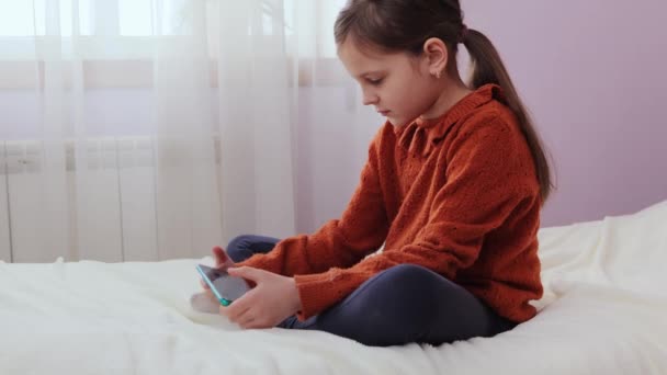デジタル幼少期 携帯電話の遊び 屋内スマートフォン利用 自宅の寝室でスマートフォンを使用してベッドに座っている驚いた少女ビデオゲーム — ストック動画