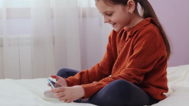 デジタルデバイスの子供 ベッドルームの技術 ワイヤレス通信プレー 自宅のベッドルームでオンラインゲームをするために携帯電話を使用してベッドに座っている楽しい少女を笑う — ストック動画
