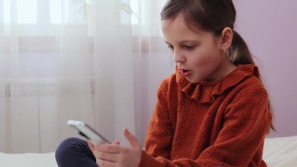 子供のデジタルワールド スマートフォンレジャー 自宅の寝室で携帯電話を使用してベッドに座っている驚いた少女は インターネットで子供のコンテンツに衝撃を与えました — ストック動画