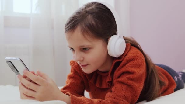 衝撃を受けた小さな茶色の毛髪の少女は オンラインスクロールや音楽を聴くために携帯電話を使用してベッドに横たわり 驚きを表現する驚くべきコンテンツを見ます — ストック動画
