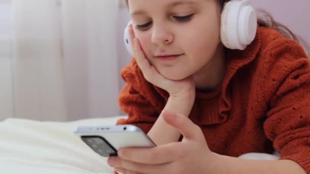 チャーミングな小さな茶色の毛髪の少女は オンラインブラウジングを楽しんでいるインターネットテクノロジーを見ながらリラックスしてスマートフォンを使用してベッドに横たわっています — ストック動画