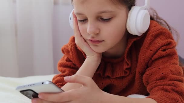 疲れた睡眠の小さな茶色のヘッドホンの少女は 携帯電話を使用してベッドに横たわり インターネットを閲覧して長時間の疲れを感じます ディスプレイを見ることから — ストック動画
