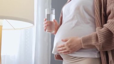 Sağlıklı hamilelik. Tanımlanamayan hamile bir kadın beyaz tişört giyip pencerenin yanında durup karnını okşayarak taze içeceğin tadını çıkarıyor.