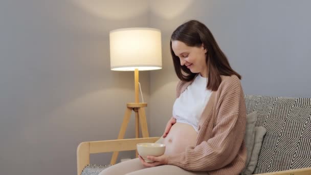 母亲的有机水果选择 怀孕期间注意营养 漂亮的棕色头发的孕妇 赤身裸体坐在沙发上 手里拿着装有新鲜水果的碗 — 图库视频影像