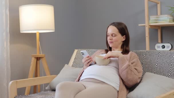 美しい妊娠中の若い女性は 将来のママのためのインターネットフォーラムを読んで自宅でソファーに座って果物を食べながら 彼女の携帯電話を使用しています — ストック動画