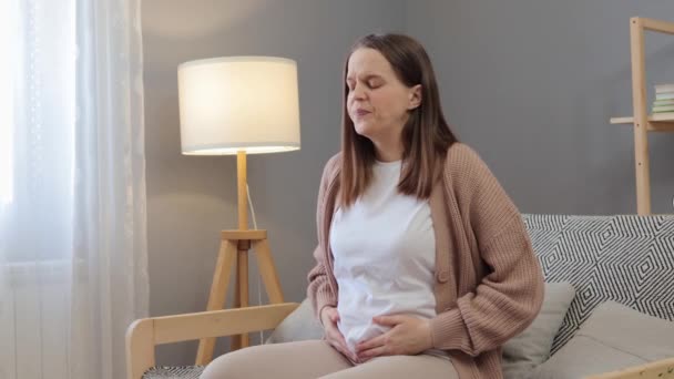 年轻的棕色头发的孕妇在家里感觉不舒服 肚子痛 在客厅里坐在沙发上 面带微笑 痛苦不堪 — 图库视频影像