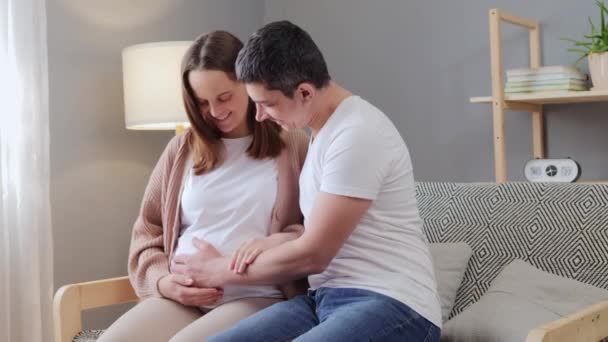 快乐的白人男人在家里拥抱孕妇 而坐在客厅的沙发上 未来的父亲正在抚摸妻子的肚子 等待孩子出生 — 图库视频影像