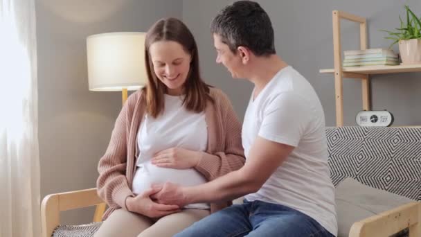 温暖的未来父母关系黑发男人和他怀孕的妻子一起坐在沙发上享受着交流和爱抚小两口的感觉 — 图库视频影像