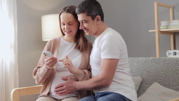 ハッピー妊娠中のカップルは リビングルームでソファに座っている彼女の夫ベビールームのインテリアを示す女性を期待して自宅で一緒に携帯電話で携帯アプリケーションを使用しています — ストック動画