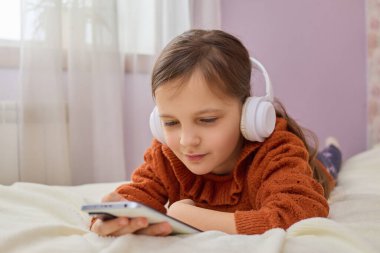 Çocuğun internet içeriği. Kafkasyalı küçük kız akıllı telefon kullanıyor mobil uygulama kullanıyor kulaklıkla müzik dinliyor ve evinde yatağında uzanmış çizgi film seyrediyor.