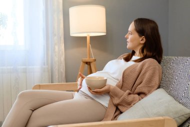 Hamilelik, beslenme sevinci. Rahat hamile kadın karnına dokunuyor kanepeye oturmuş taze meyveyle oturmuş pencereye bakıyor ve organik atıştırmalıkların tadını çıkarıyor.