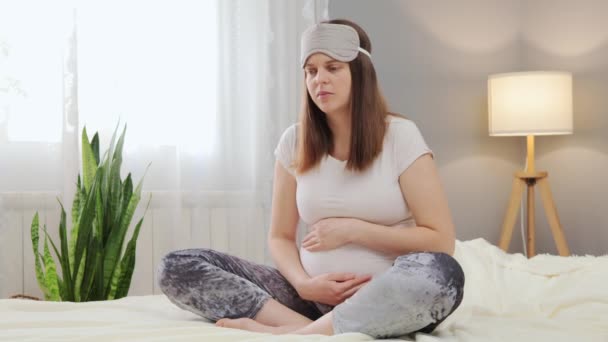 不健康な妊婦が自宅の内装でベッドに座り 額に寝たマスクを着けながら 不快感や腹痛を感じている — ストック動画